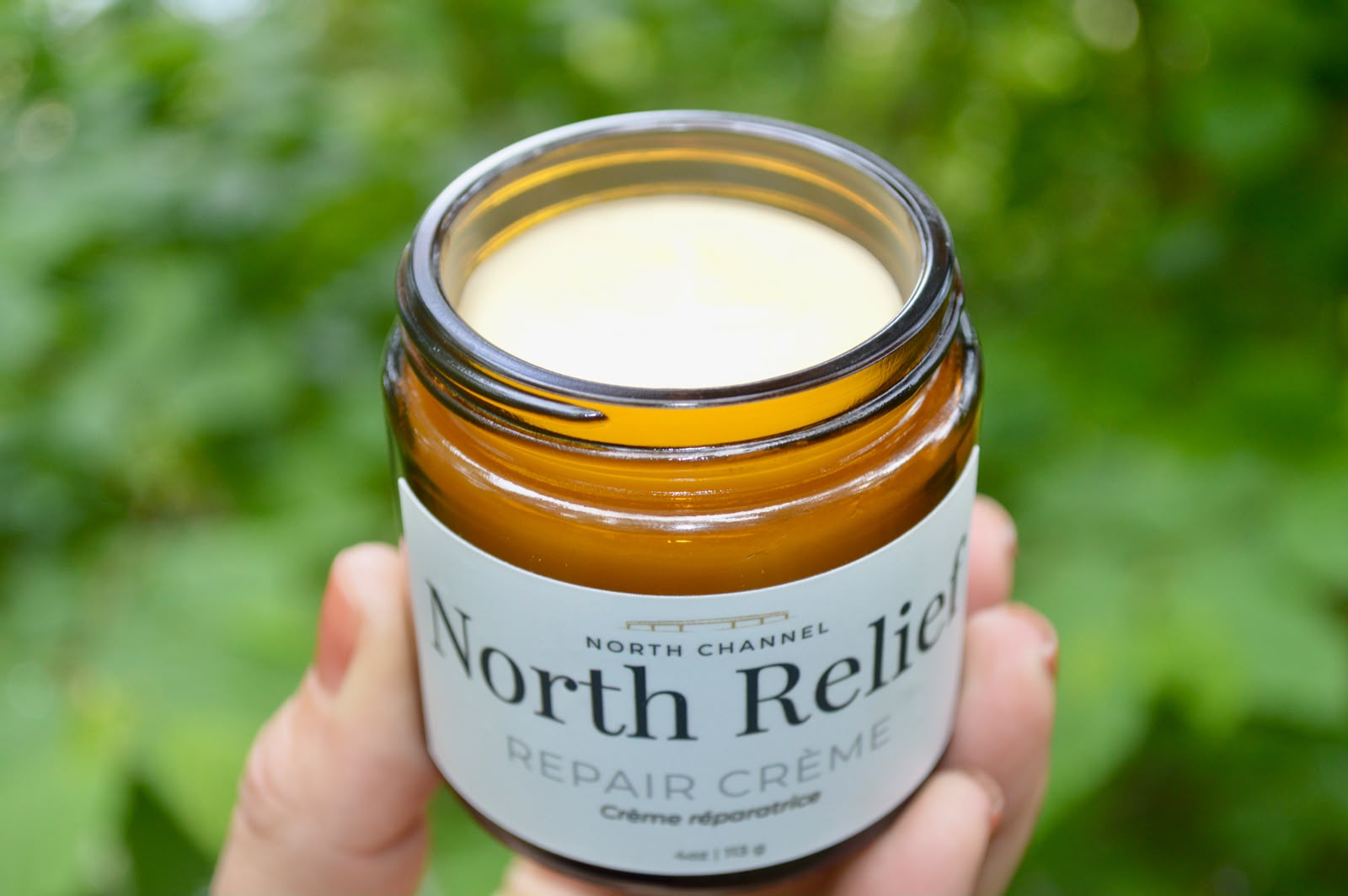 An open jar of North Relief repair crème held in one hand, green Northern foliage behind